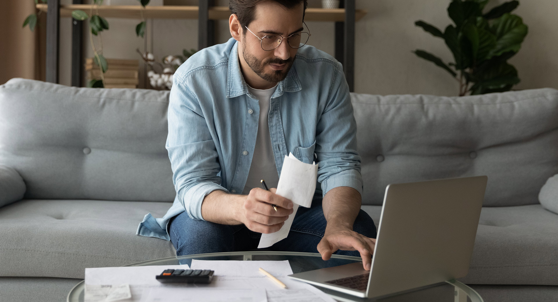 A person sitting on a sofa holds a pen and paper in one hand and types on a laptop keyboard with the other