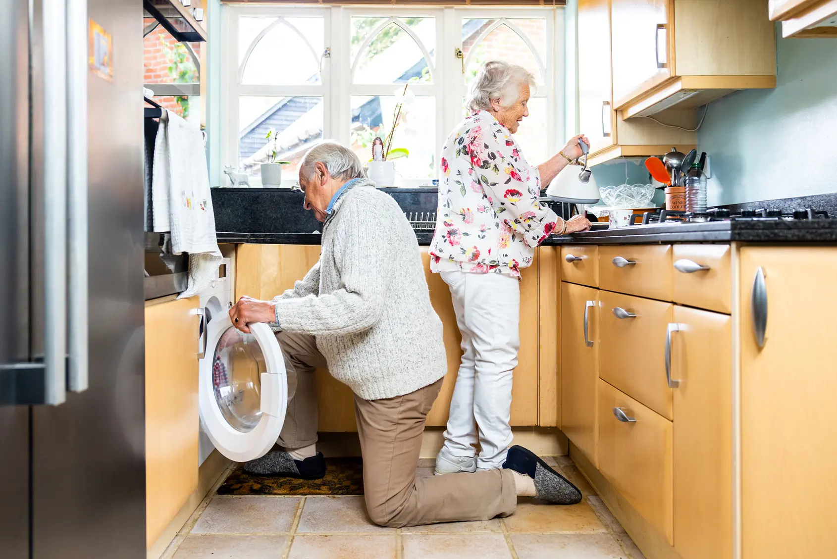 An elderly couple in their kitchen, one prepares food while the other puts a towel into the washing machine