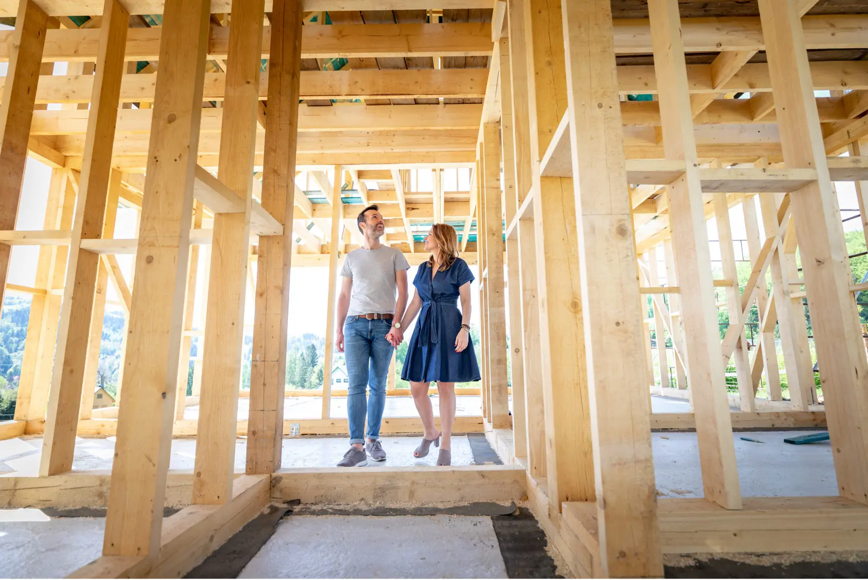 A couple smile and stand inside a timber frame of a house on a sunny day
