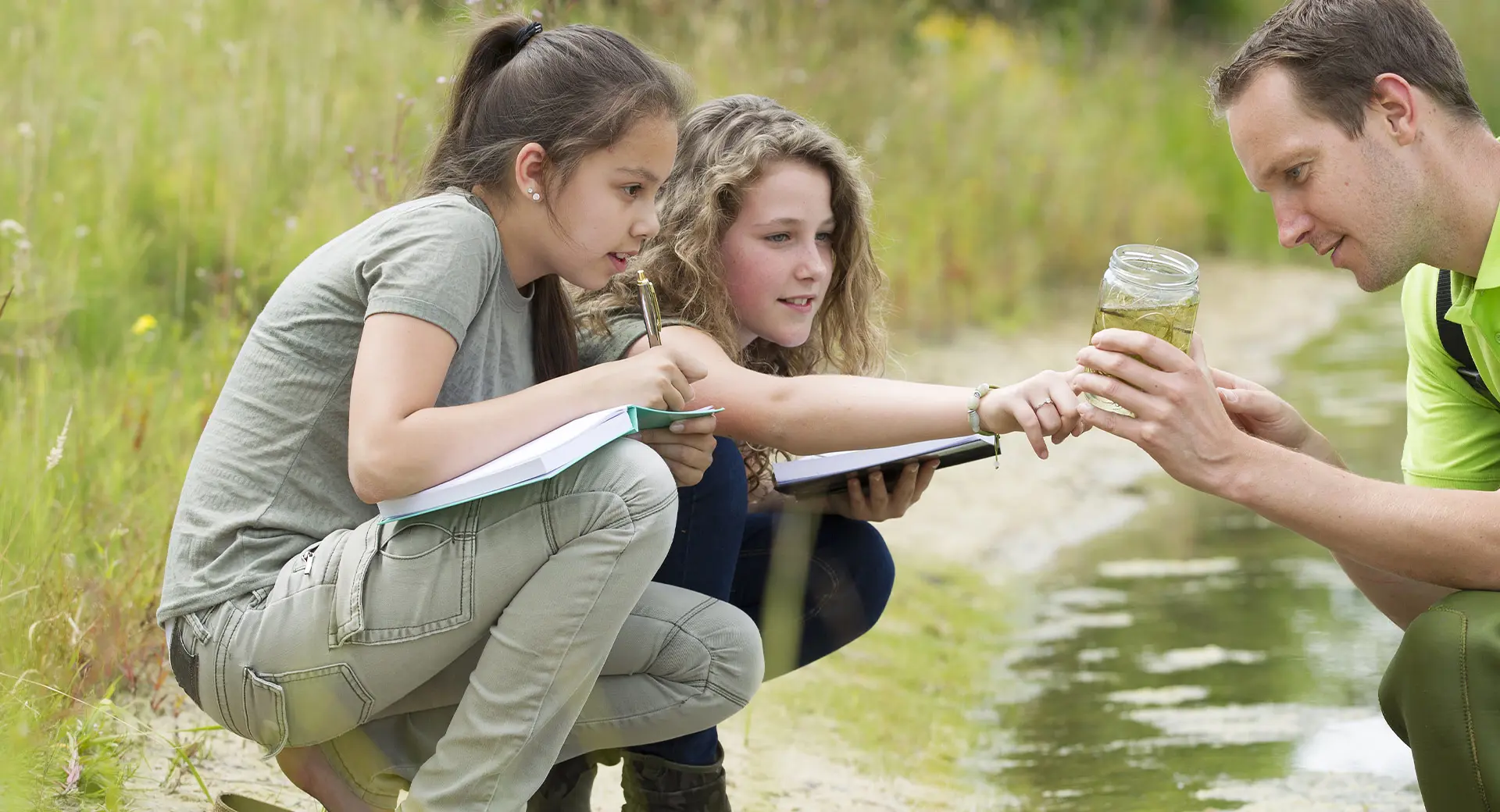 At the edge of a pond, two children and an adult inspect a jar full of pond life