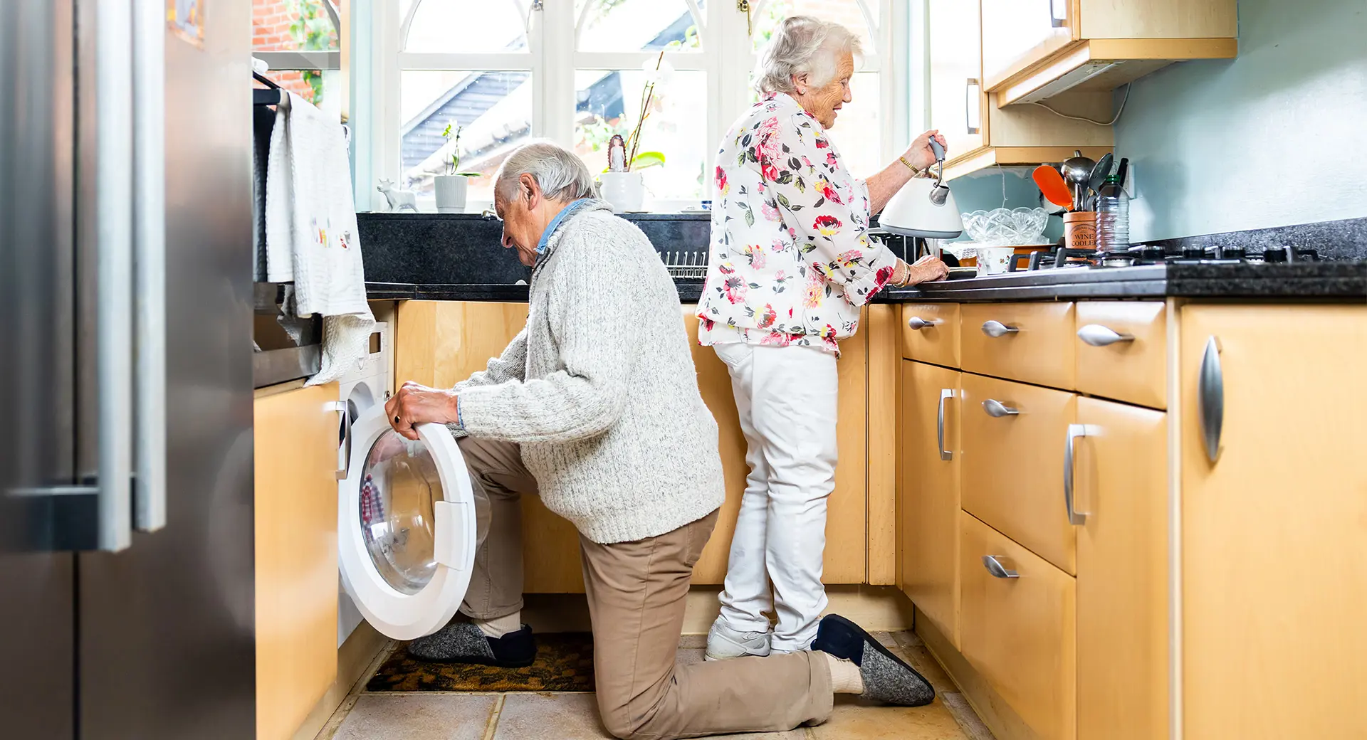 An elderly couple in their kitchen, one prepares food while the other puts a towel into the washing machine